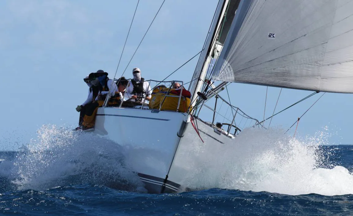 SUMMER STORM crashing upwind through a wave, spray over the bow, crew in foul-weather gear on the rail, white headsail set