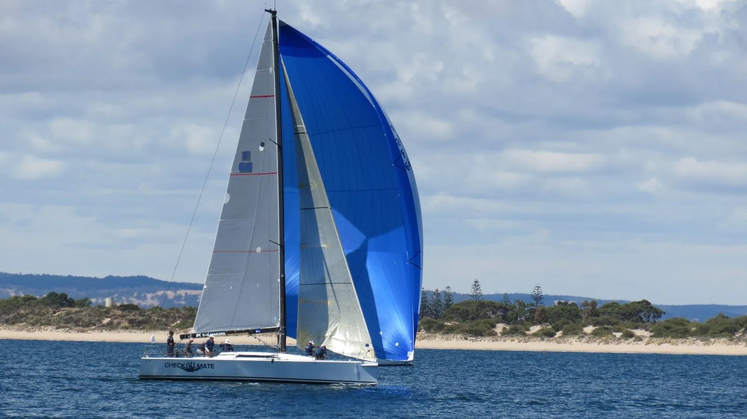 Summit 40 CHECKMATE reaching under a blue asymmetric spinnaker, white mainsail and jib along a low sandy coastline