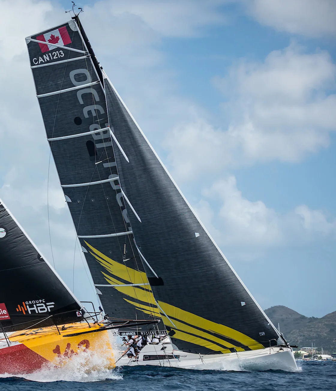 Pogo 12.5 CAN 1213 racing under a black X-Drive® mainsail with yellow wing graphics beside a yellow-hulled boat, hills astern