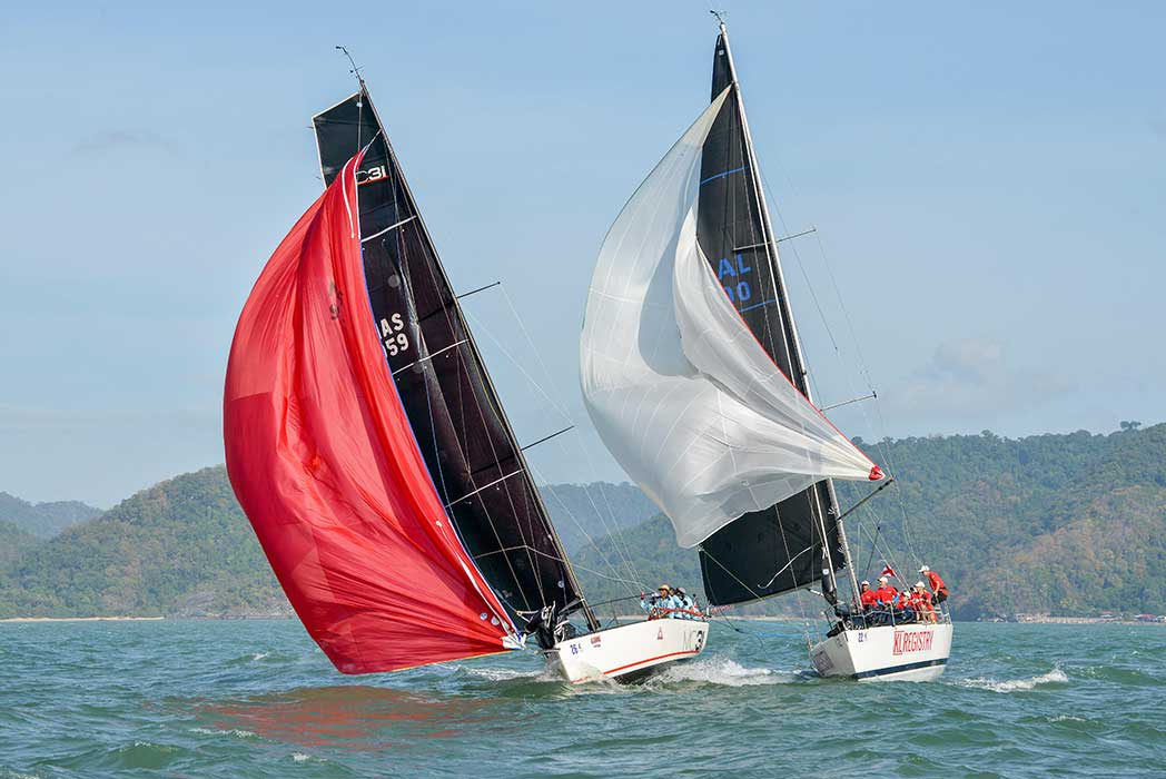 Two racing yachts overlapped downwind, one under a red spinnaker and one under a white spinnaker, hills behind