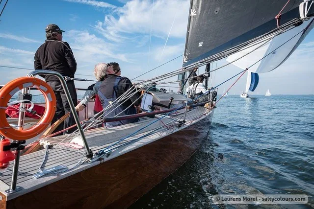 Looking forward along the Waarschip 1012's varnished deck in light air, crew seated and other yachts in the distance