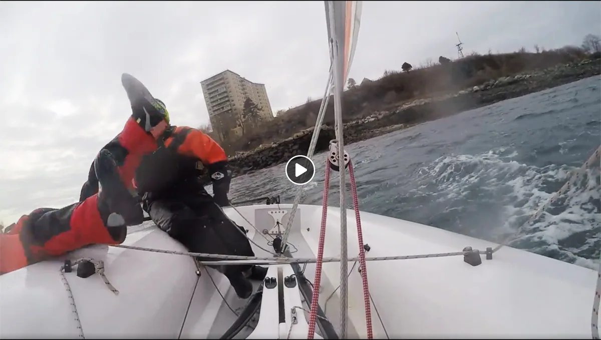 Video still from a small racing boat's deck, crew in red foul-weather gear hiking as it sails near a city shoreline