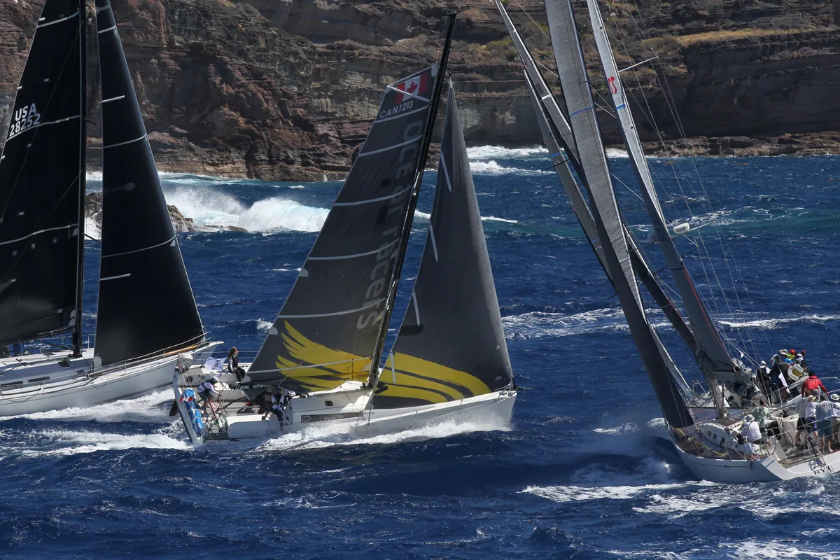 Three racing yachts on converging courses under dark sails with rocky cliffs and breaking surf behind