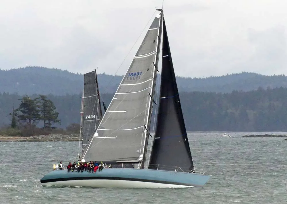 Long light blue racing sloop with grey sails and crew on the rail leads a black-sailed boat past a wooded shore
