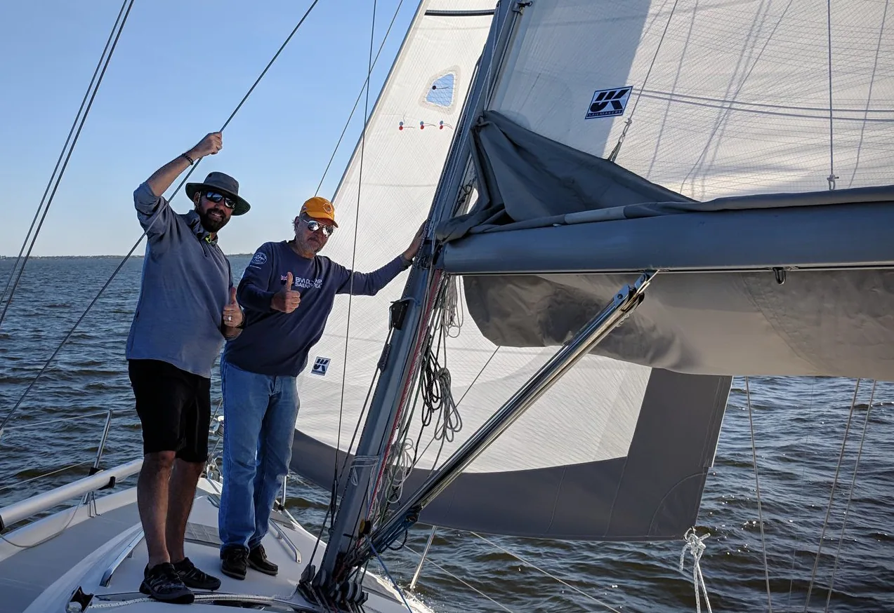 Two smiling sailors on deck, one giving a thumbs up, beside a new white mainsail with a UK Sailmakers logo