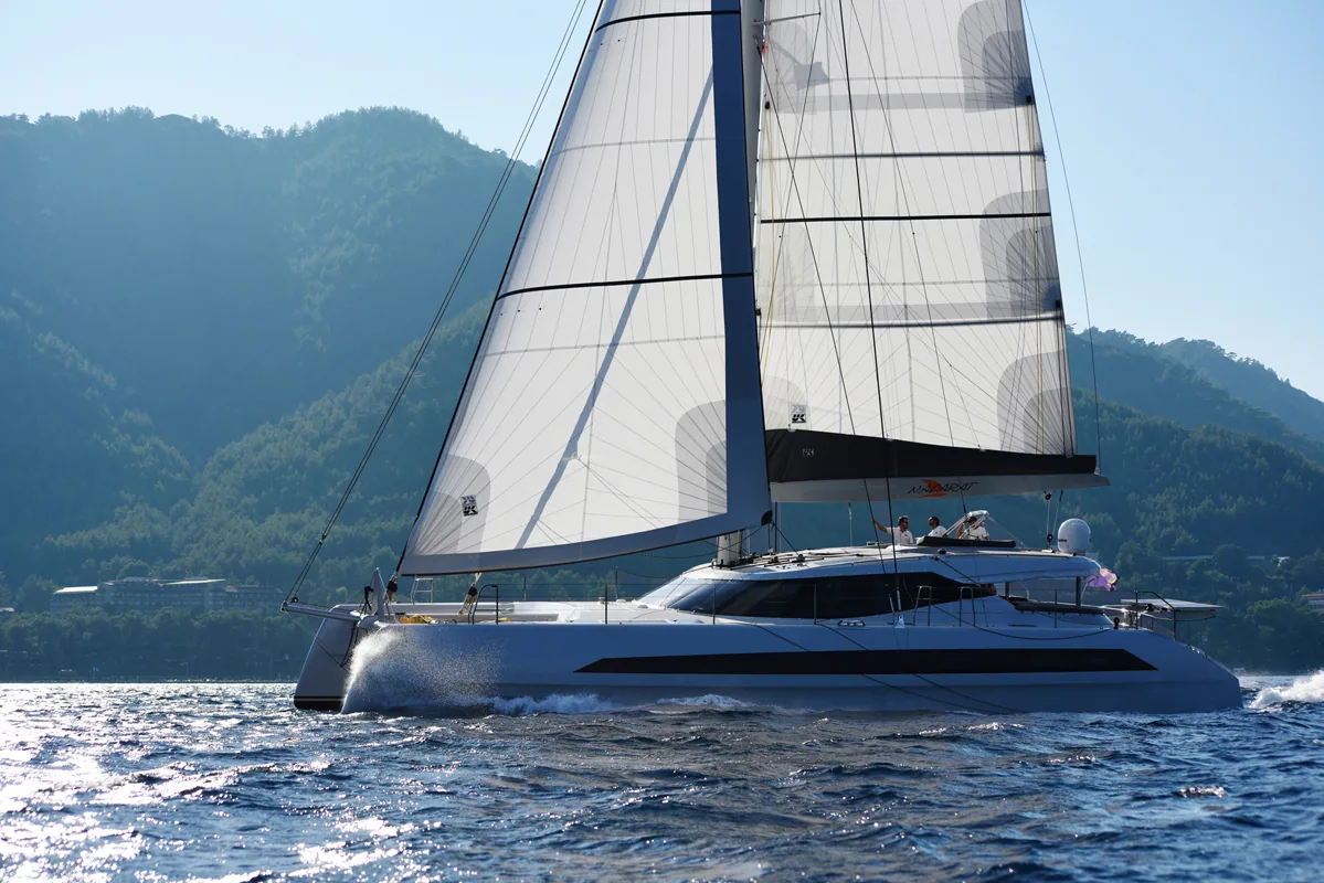 Dubois 60 catamaran sailing under a full-battened white mainsail and genoa with green mountains behind