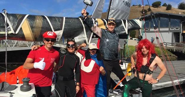 Five sailors in fancy dress, including a Mario costume and a red-wigged mermaid, posing on a dock before their boat