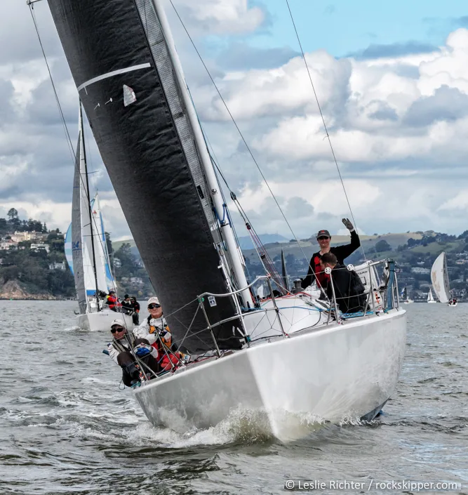 White Figaro 2 sailing upwind under a black mainsail, crew waving from the cockpit, spinnakers and hills behind