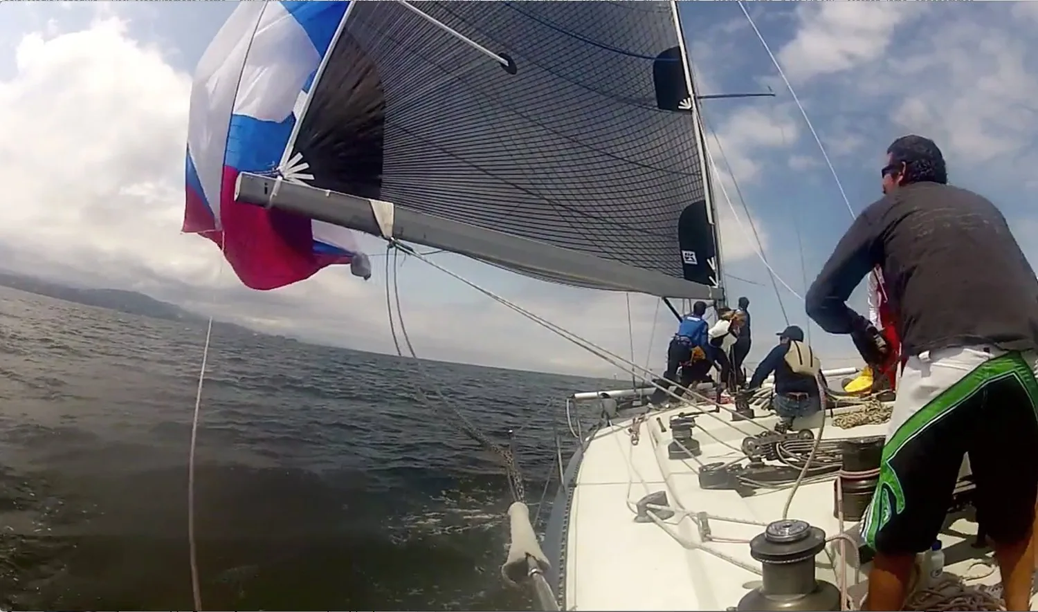 On-deck view of a red, white and blue spinnaker flogging above the bow as crew scramble forward on a racing sloop
