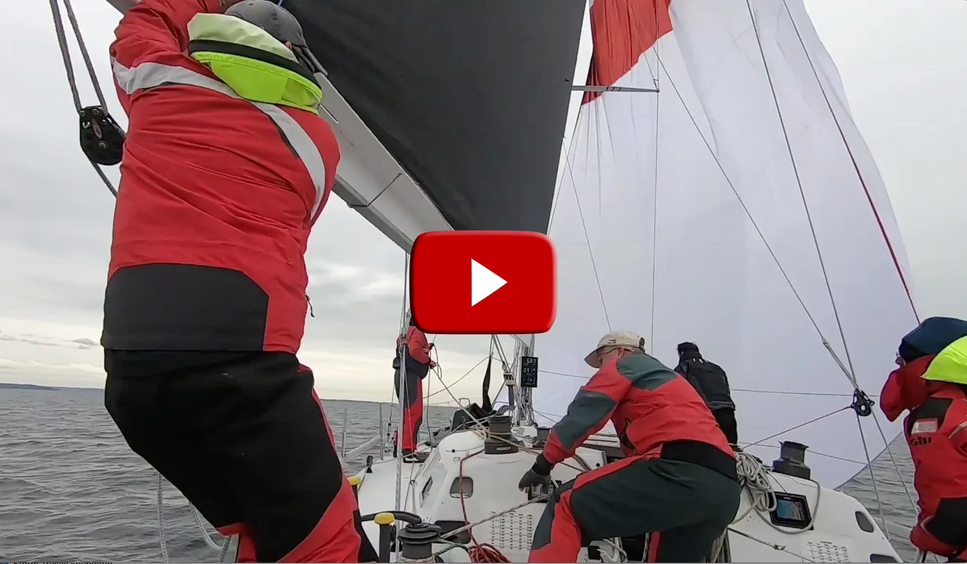 Video still of crew in red foul-weather gear working a red and white asymmetric spinnaker through a gybe