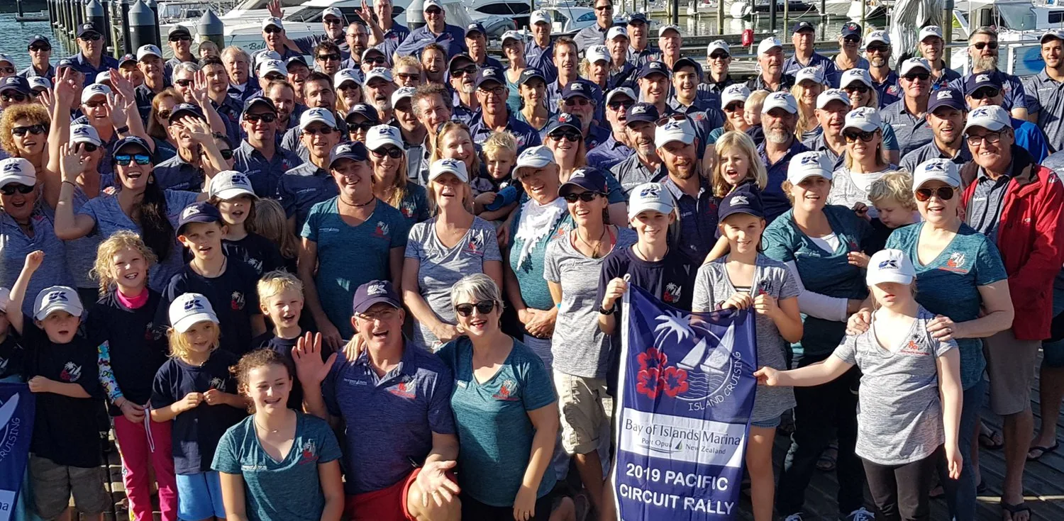 Large group of sailors and children in matching caps holding a 2019 Pacific Circuit Rally banner at a marina