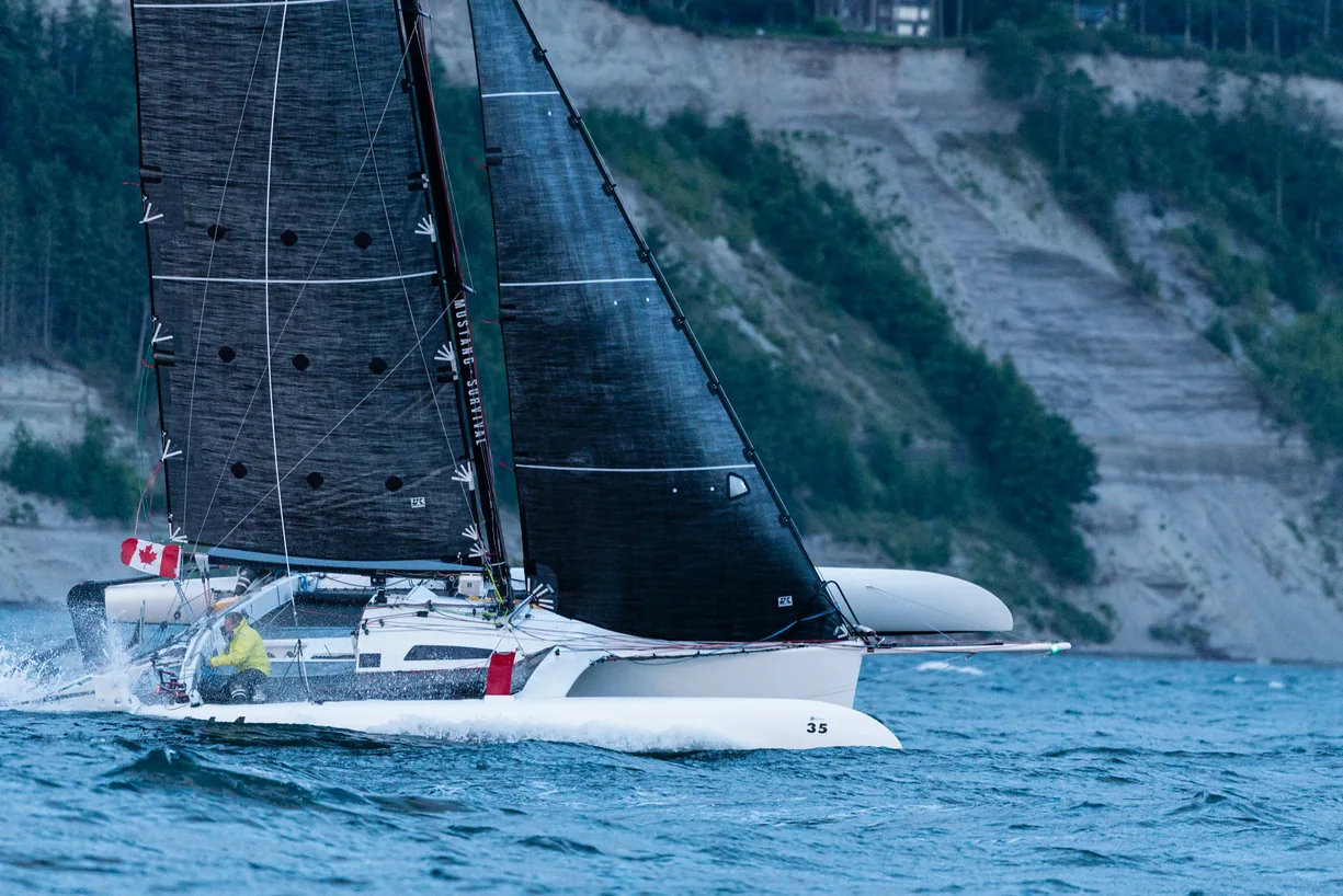 Trimaran under black UK Titanium mainsail and jib flying a Canadian flag along a cliff-lined shore in the Race to Alaska