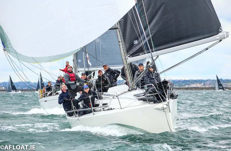 Racing yacht beating upwind under black mainsail and jib, crew hiked on the rail waving, a white spinnaker overhead nearby