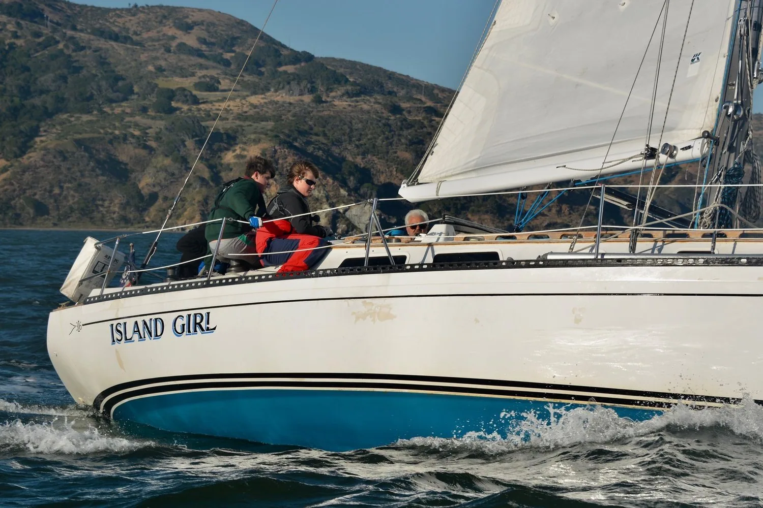 ISLAND GIRL heeled upwind with two crew on the rail, white hull with blue boot stripe, hills behind