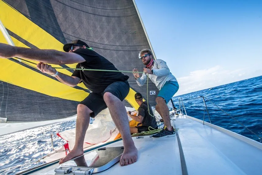 Two barefoot foredeck crew hauling a sheet under a yellow-and-black asymmetric spinnaker on a Pogo 12.50