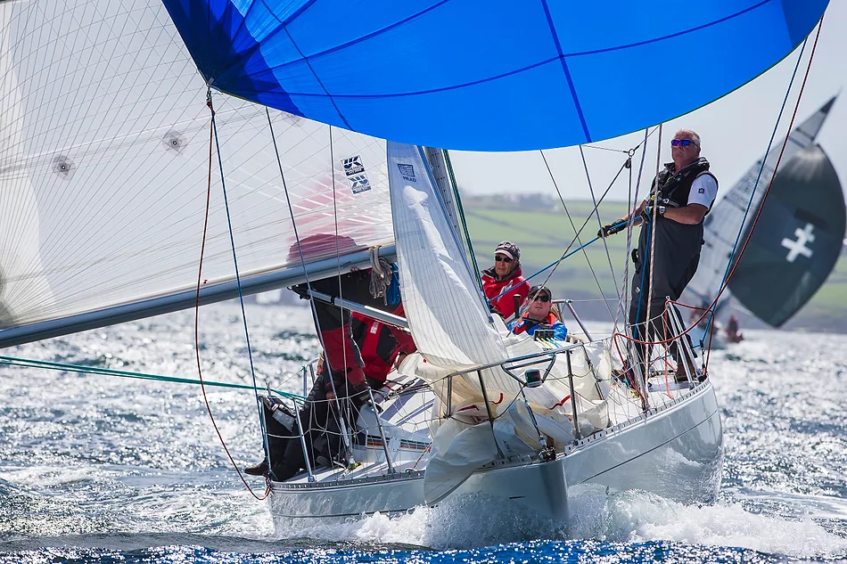 Crew in red gybing under a blue spinnaker with the jib bundled on the bow, a black-sailed rival astern under green hills