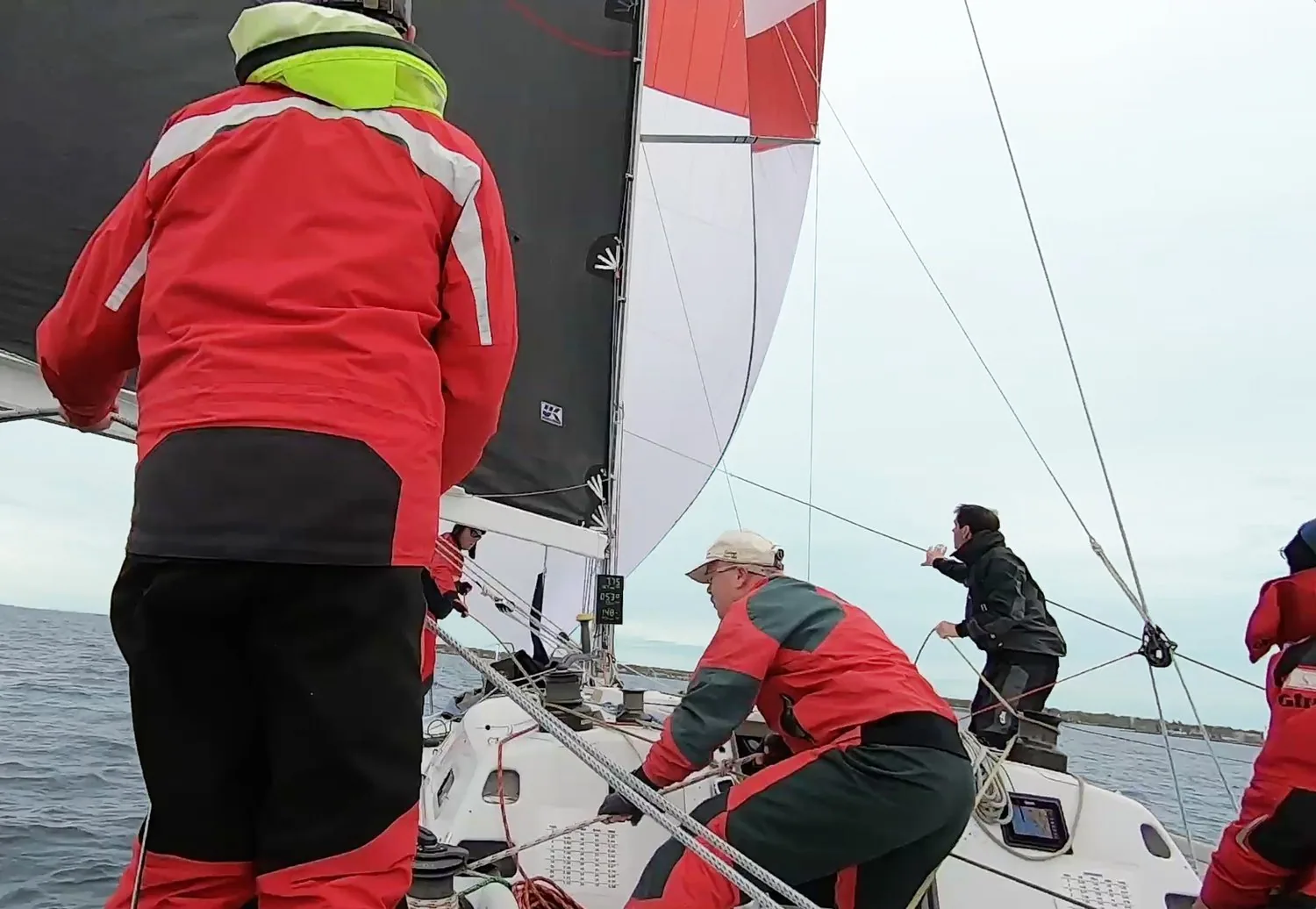 On deck mid-gybe: crew in red foul-weather gear, grey mainsail and a white spinnaker with red panels filling ahead