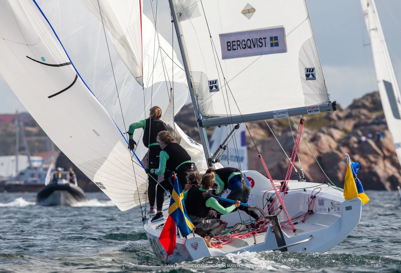 Women's match-racing crew in black hiking on a white keelboat with a BERGQVIST boom banner and yellow flag off a rocky shore
