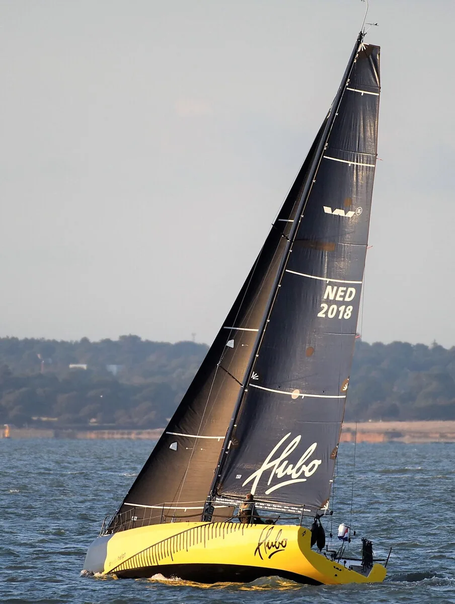 HUBO, NED 2018, seen from astern beating upwind under black mainsail and jib, yellow hull, tree-lined shore behind