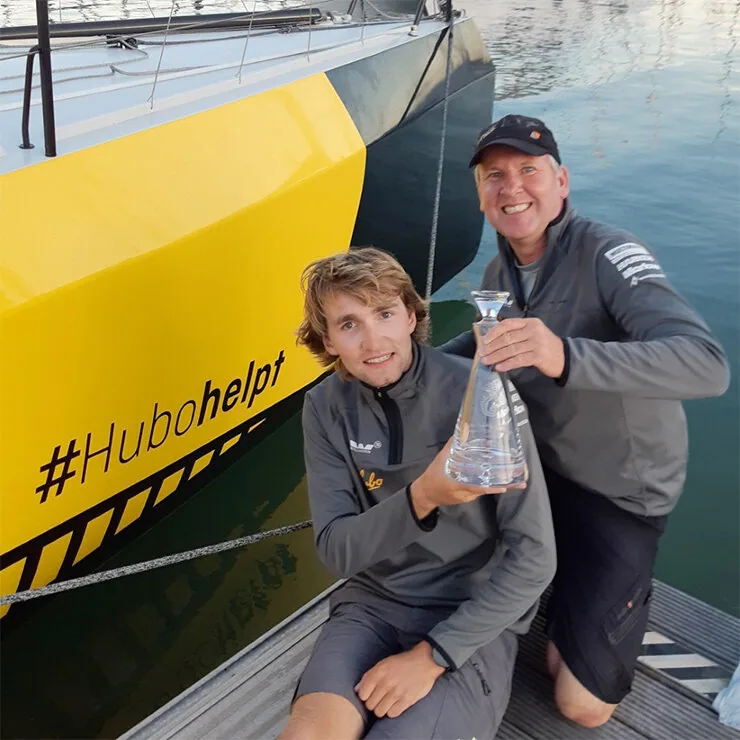 Jochem Schoorl and Eric van Vuuren holding a glass trophy on the dock beside HUBO's yellow hull marked #Hubohelpt