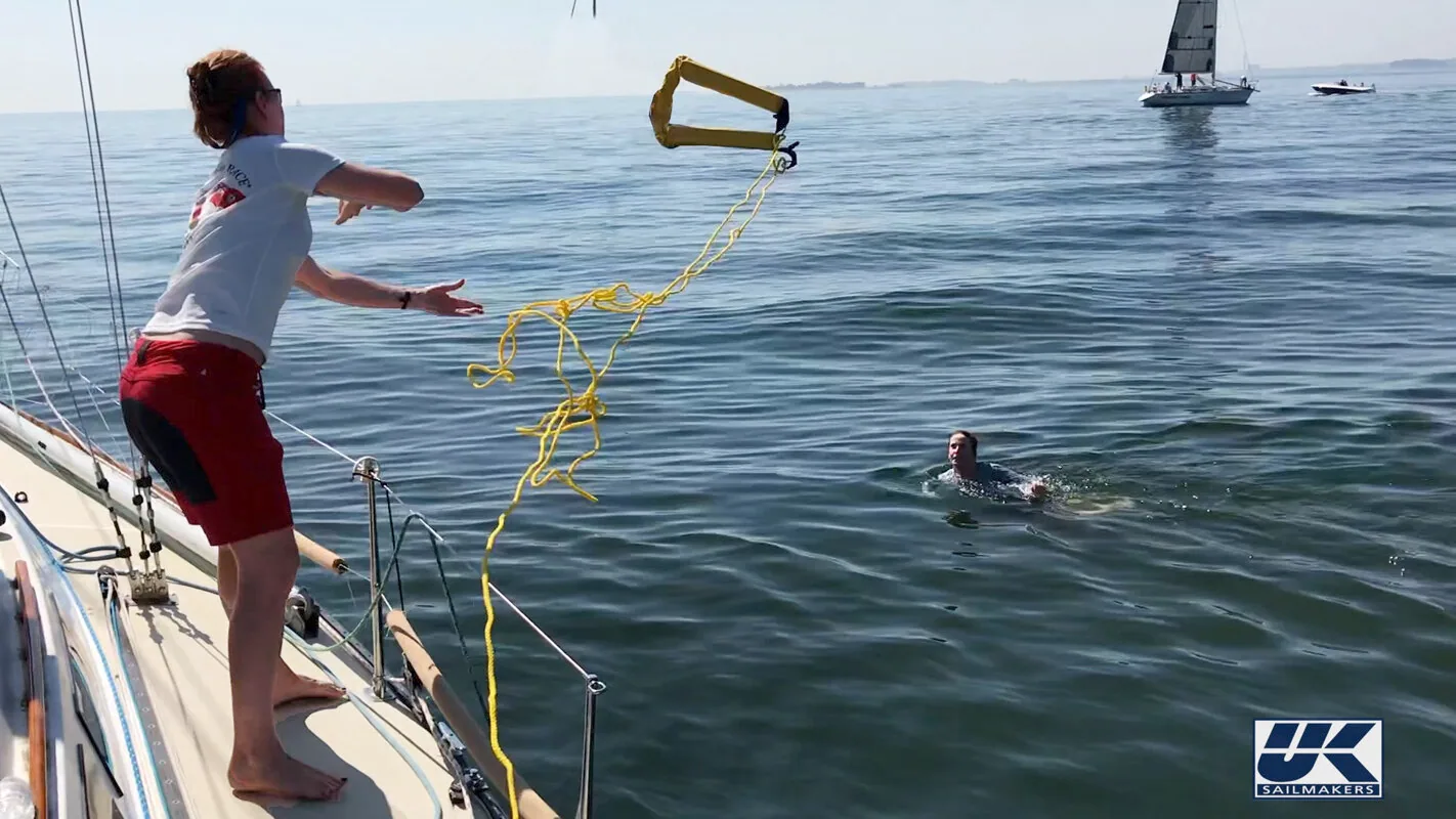 Crew member on deck throwing a yellow LifeSling and floating line toward a swimmer in calm water