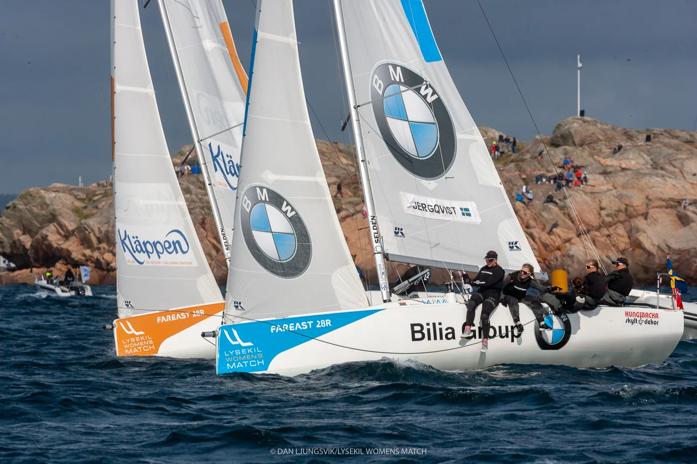 Two match-racing keelboats with BMW logos on their mainsails duelling close together off a rocky shore lined with spectators