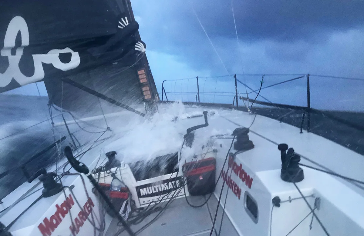 Waves sweeping across HUBO's cockpit under a stormy sky, black mainsail with Hubo script overhead, Marlow and Harken stickers