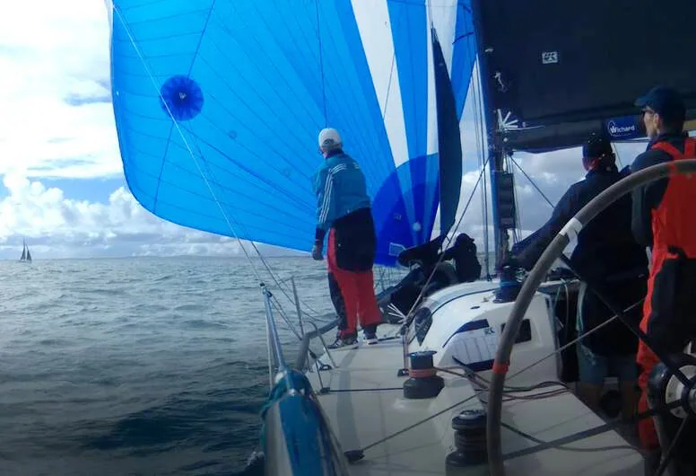 Onboard view sailing wing-on-wing: blue spinnaker set to windward, dark mainsail to leeward, crew at the mast and helm