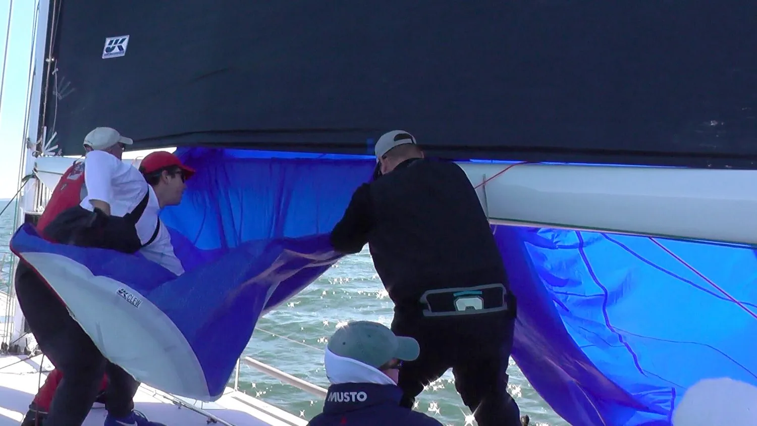 Crew hauling a blue spinnaker down through the letterbox slot between the boom and the foot of a black UK Sailmakers mainsail