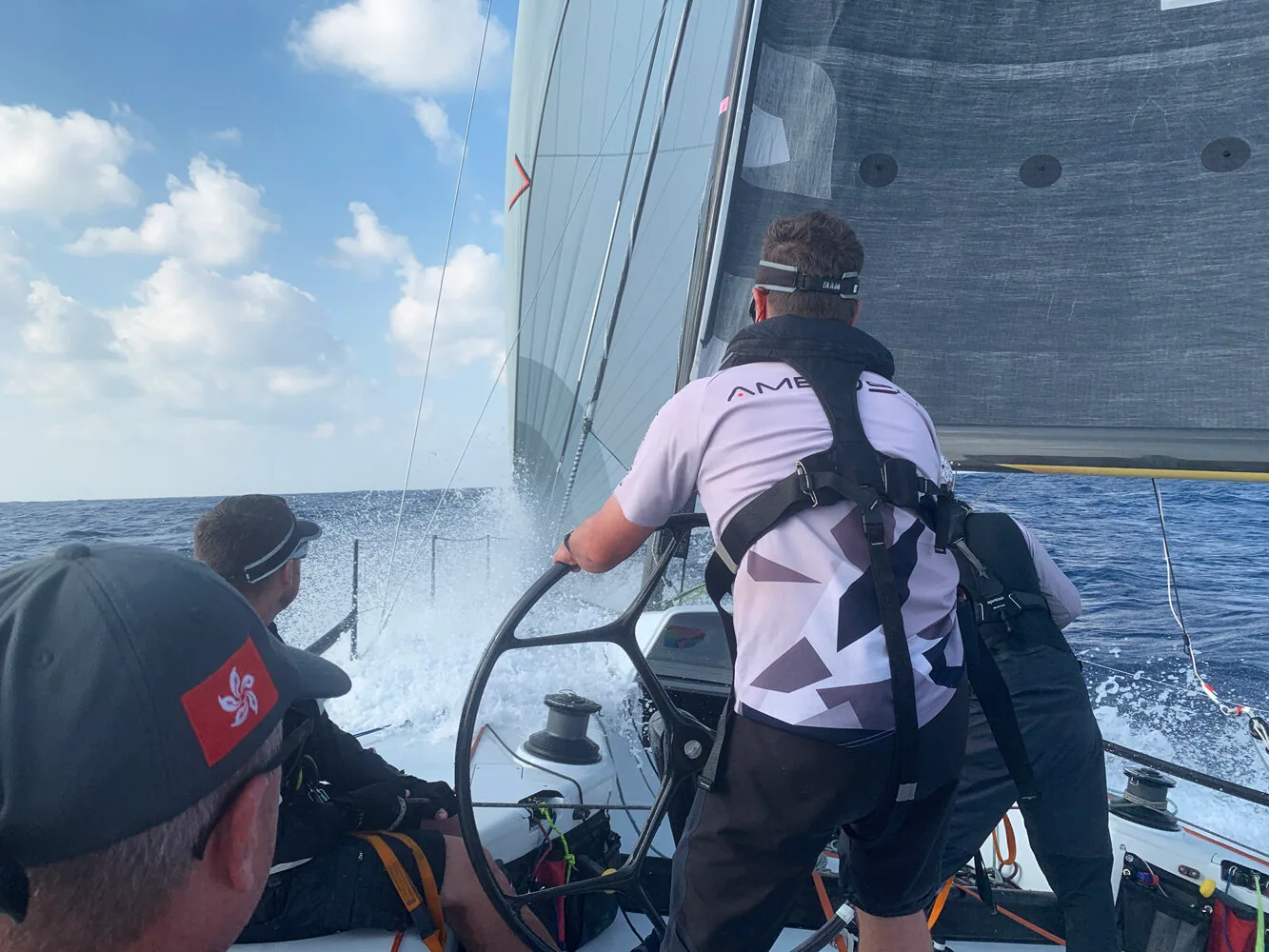 Helmsman in an AMBUSH crew shirt at the wheel as a wave breaks over the bow, grey mainsail and jib overhead