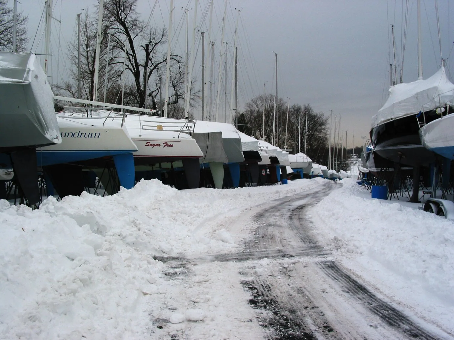 Sailboats under winter covers on stands in a snow-buried boatyard, a plowed track running between the rows