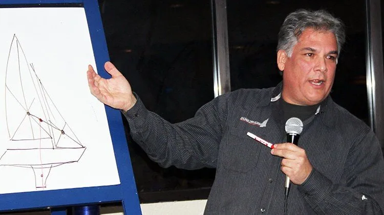 Juan Mauri holding a microphone and gesturing at a hand-drawn sail-plan sketch on a whiteboard