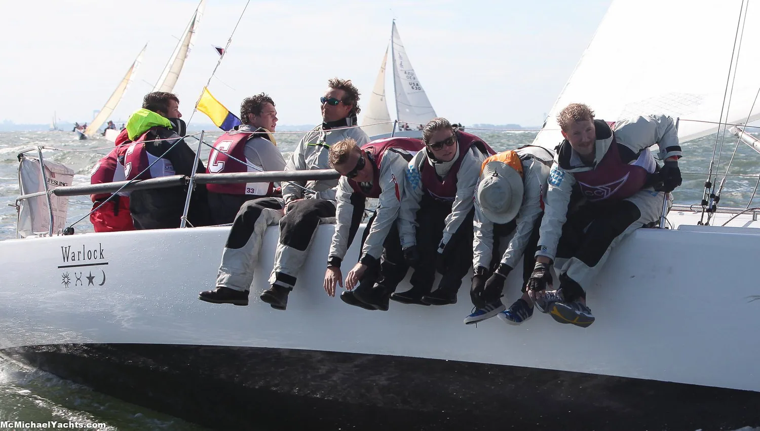 Crew of the sloop WARLOCK hiking hard on the weather rail upwind, spinnakers of other boats astern