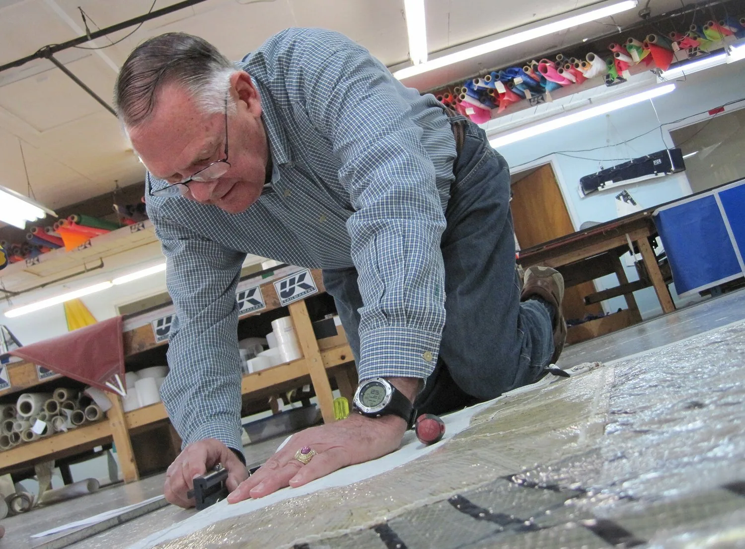 Butch Ulmer kneeling on the loft floor in a checked shirt, hand-finishing a laminate sail panel