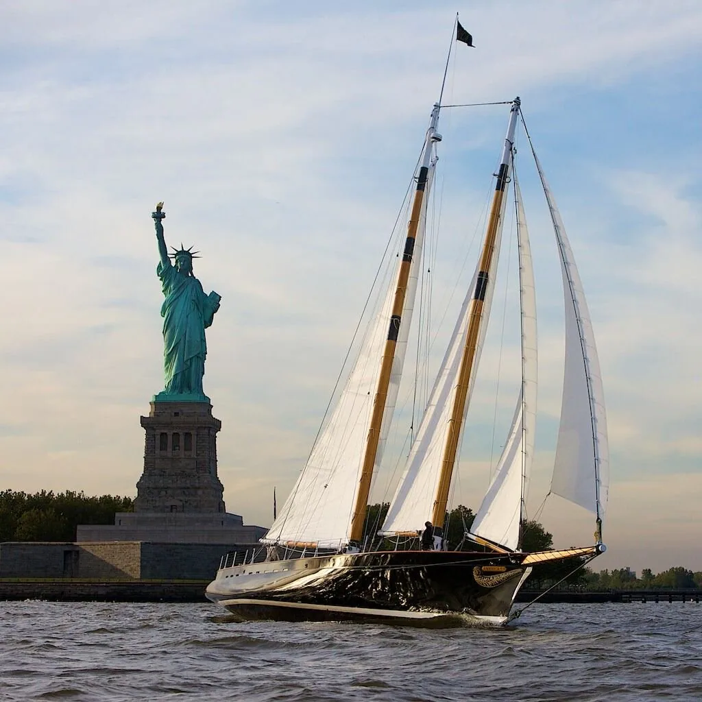 Black-hulled schooner AMERICA 2.0 under full white sail passing the Statue of Liberty
