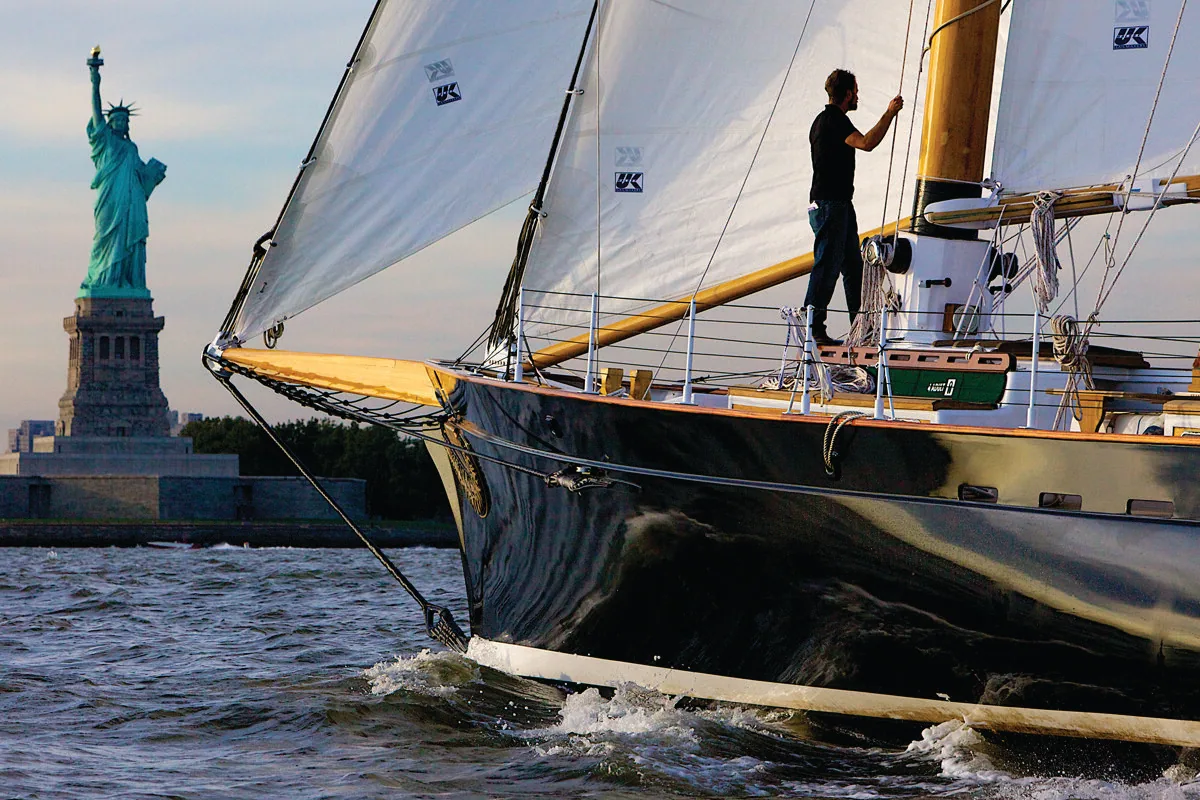 Bow of the schooner AMERICA 2.0 with UK Sailmakers logos on her white sails, a crewman aboard, Statue of Liberty astern