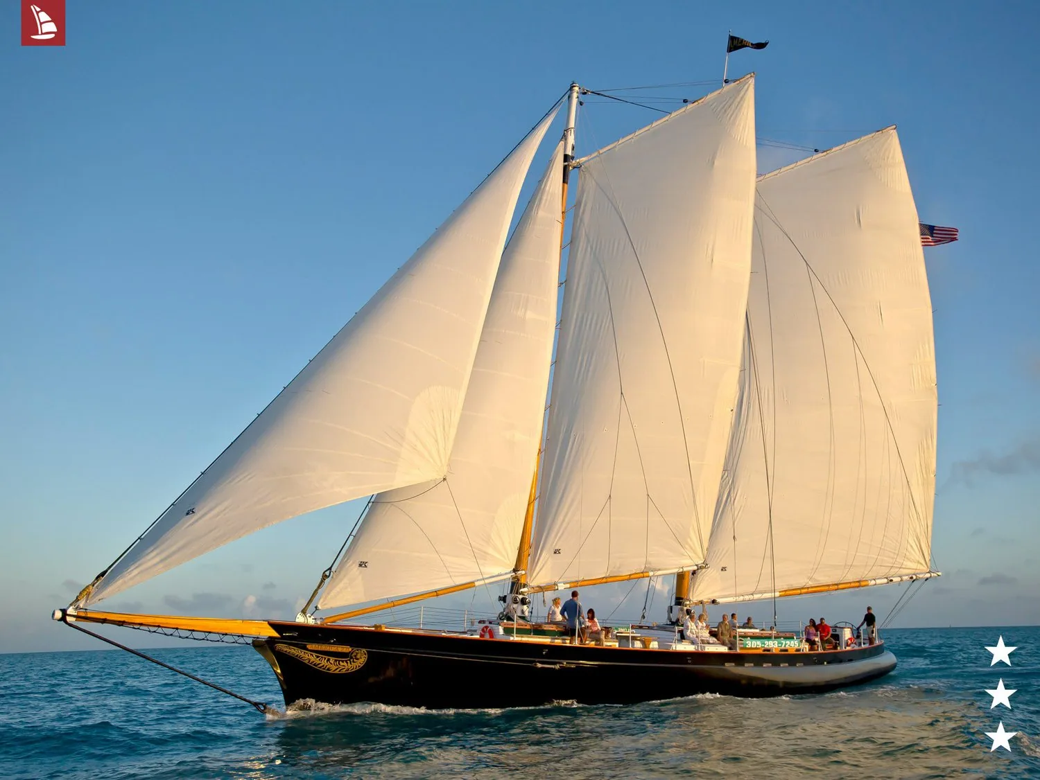 Schooner AMERICA 2.0 under full sail in golden evening light with passengers along the rail