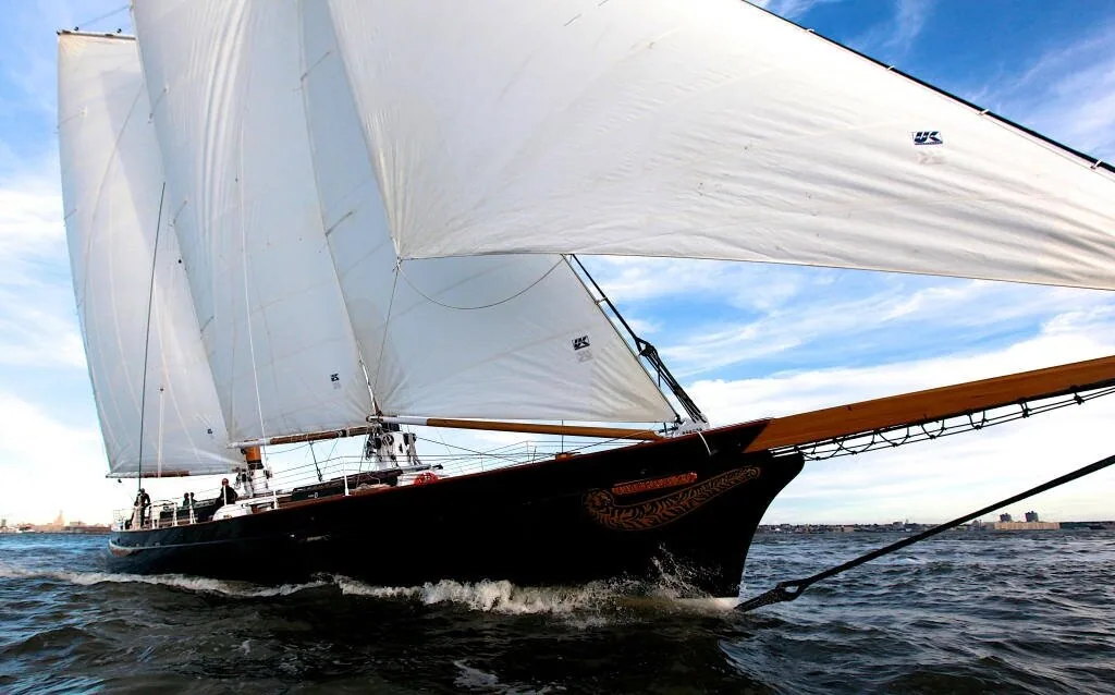 Low bow-on view of the black schooner AMERICA 2.0 heeling under white sails, bowsprit reaching over the water