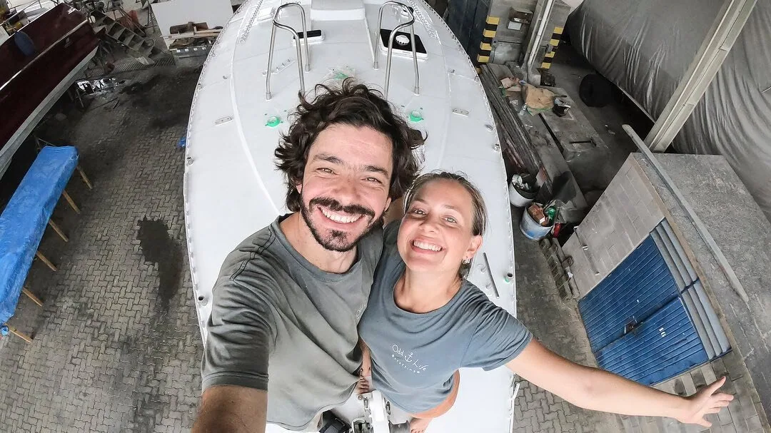 The Odd Life Crafting couple grinning in a selfie beside the white hull of their boat in a workshop