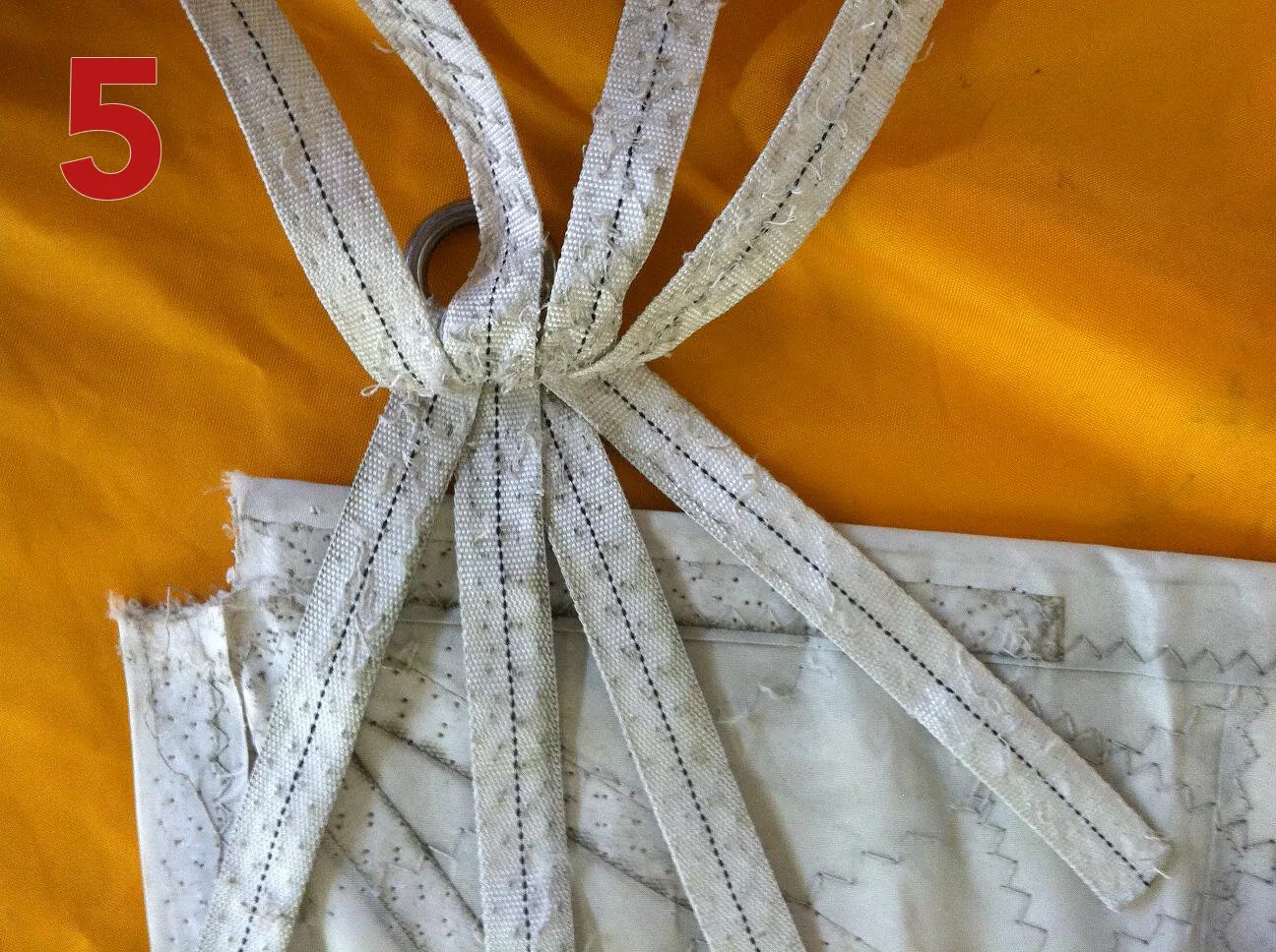 Frayed and UV-degraded white webbing straps at a sail's clew ring, laid on orange cloth, labelled 5