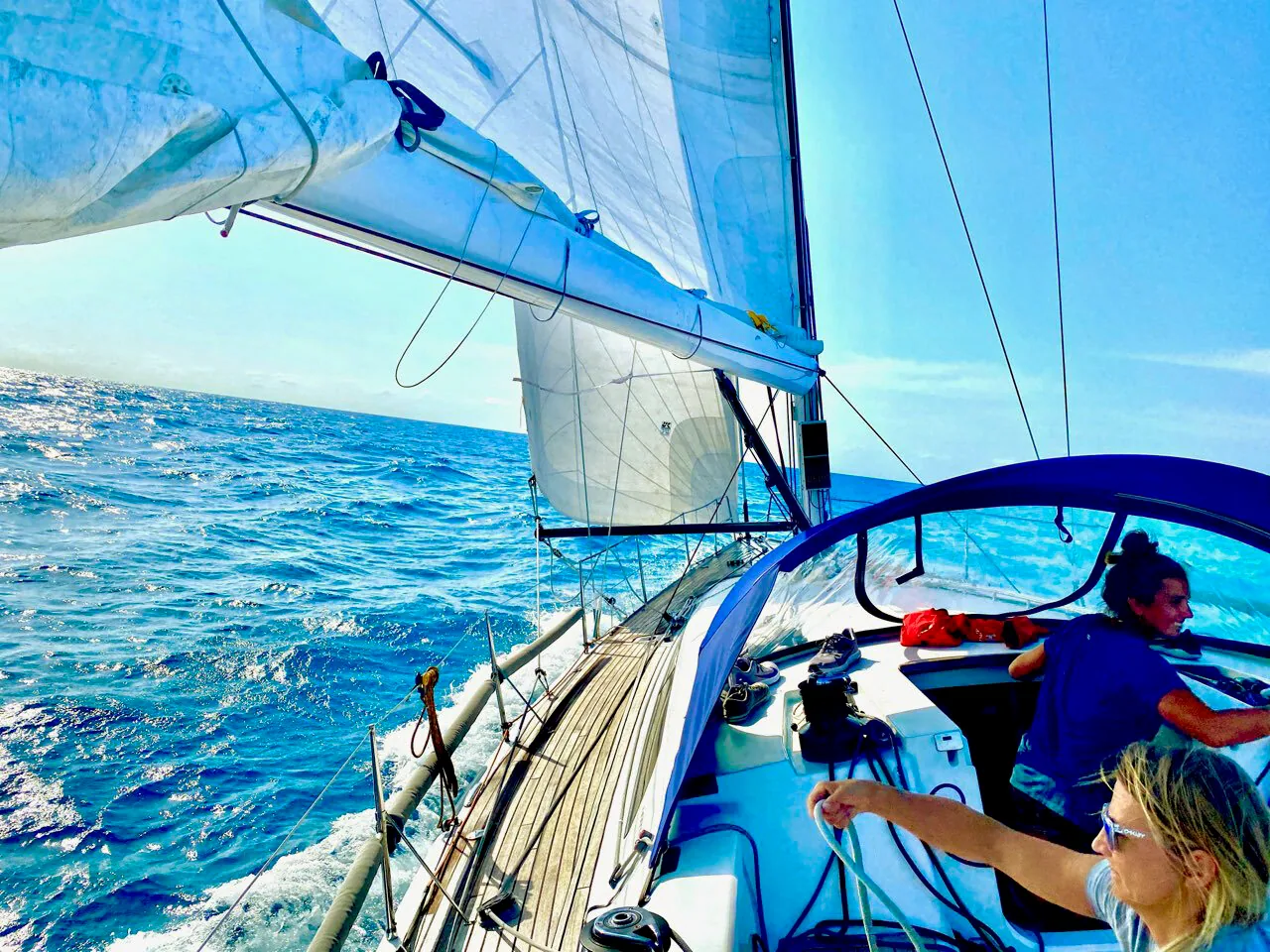 Sailboat reaching in bright sun with the jib eased, two crew relaxed in the cockpit behind a teak side deck