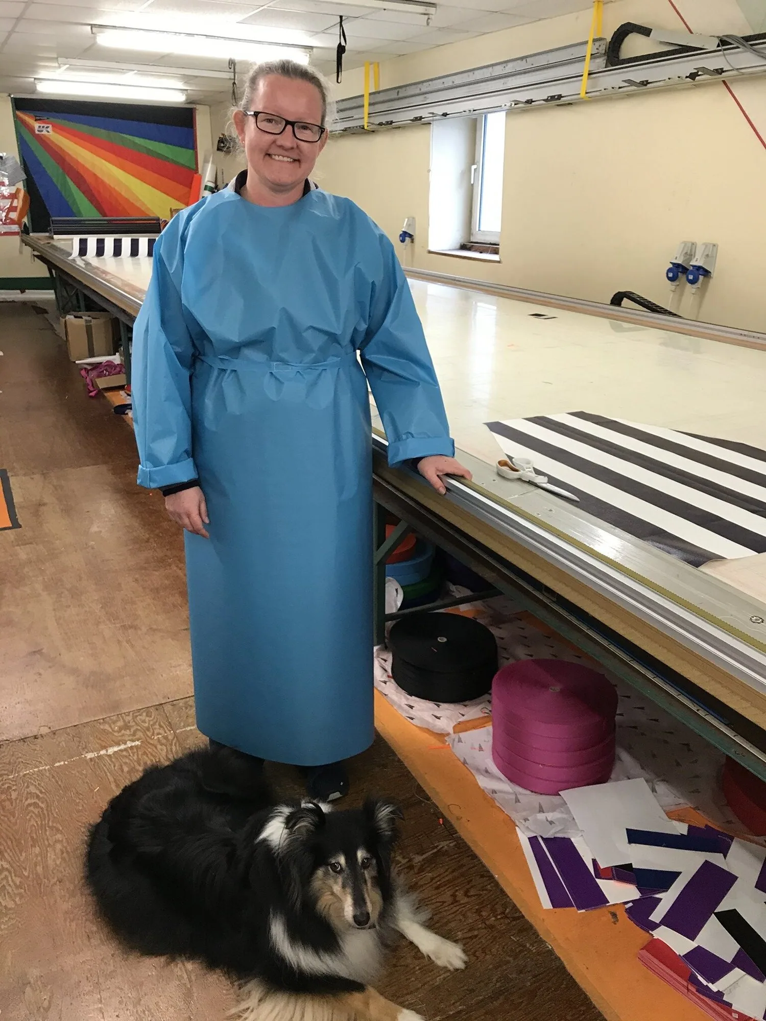 Woman in glasses wearing a blue isolation gown beside a sail loft cutting table, a collie lying at her feet