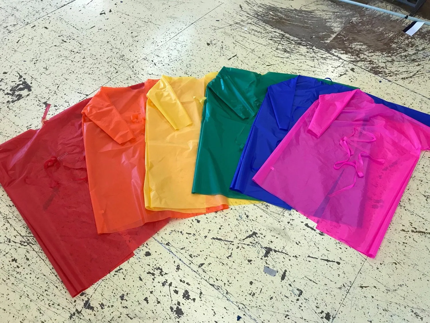 Six isolation gowns in red, orange, yellow, green, blue and pink laid out on a paint-spattered loft floor