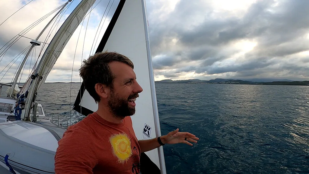 Smiling bearded sailor in an orange T-shirt on deck under grey skies, white headsail and a coastline behind