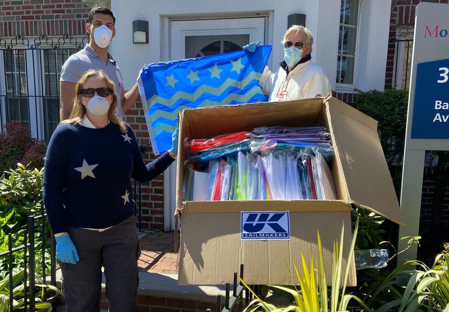 Three masked volunteers holding up a UK Sailmakers box of colourful sailcloth PPE outside a brick building