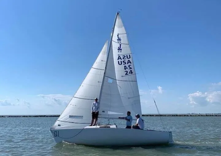 J/22 with white main and jib, sail number USA 24, three crew aboard on flat water under a blue sky