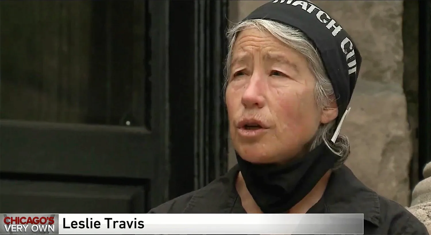 TV news frame of Leslie Travis, grey-haired with a black headband and pulled-down face mask, name caption below