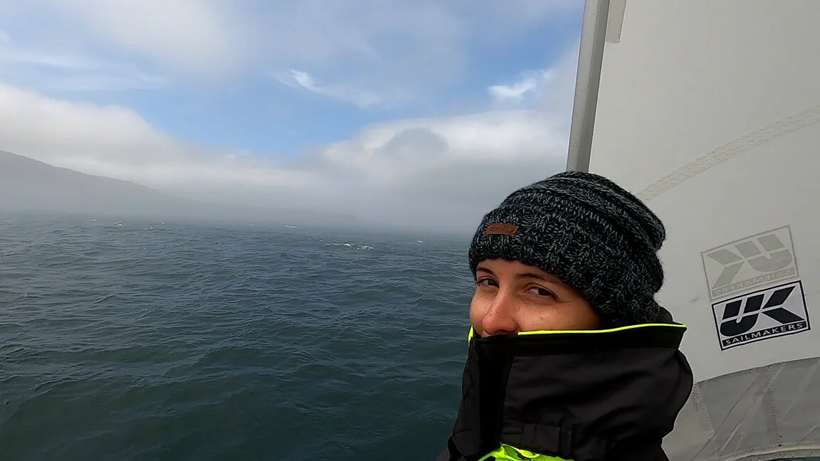 Sailor in a knit beanie and zipped jacket at the rail as fog lifts off a Maine hillside, UK Sailmakers sail behind
