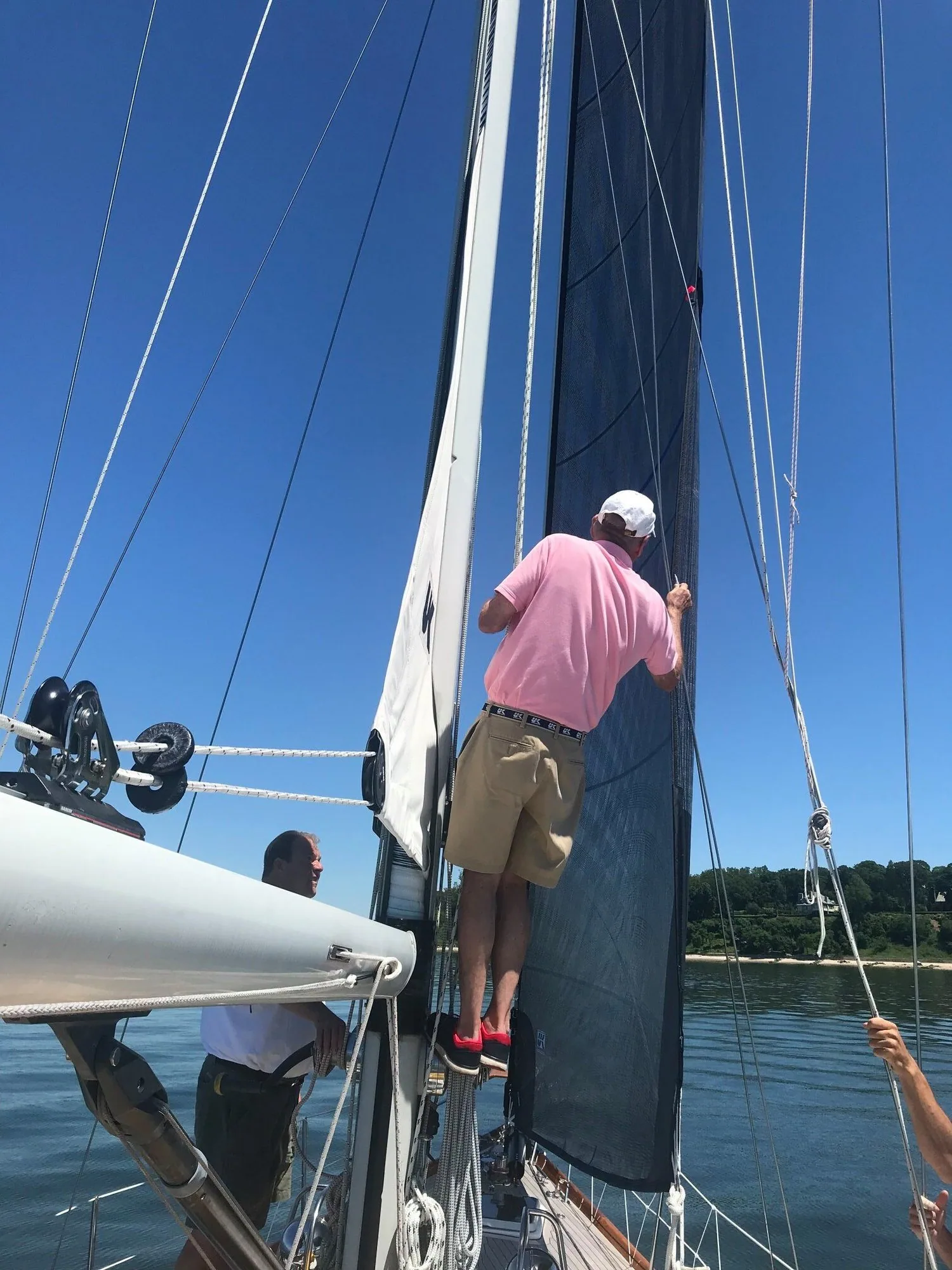 Sailmaker in a pink polo and cap on deck inspecting the luff of a dark blue headsail, another crew watching