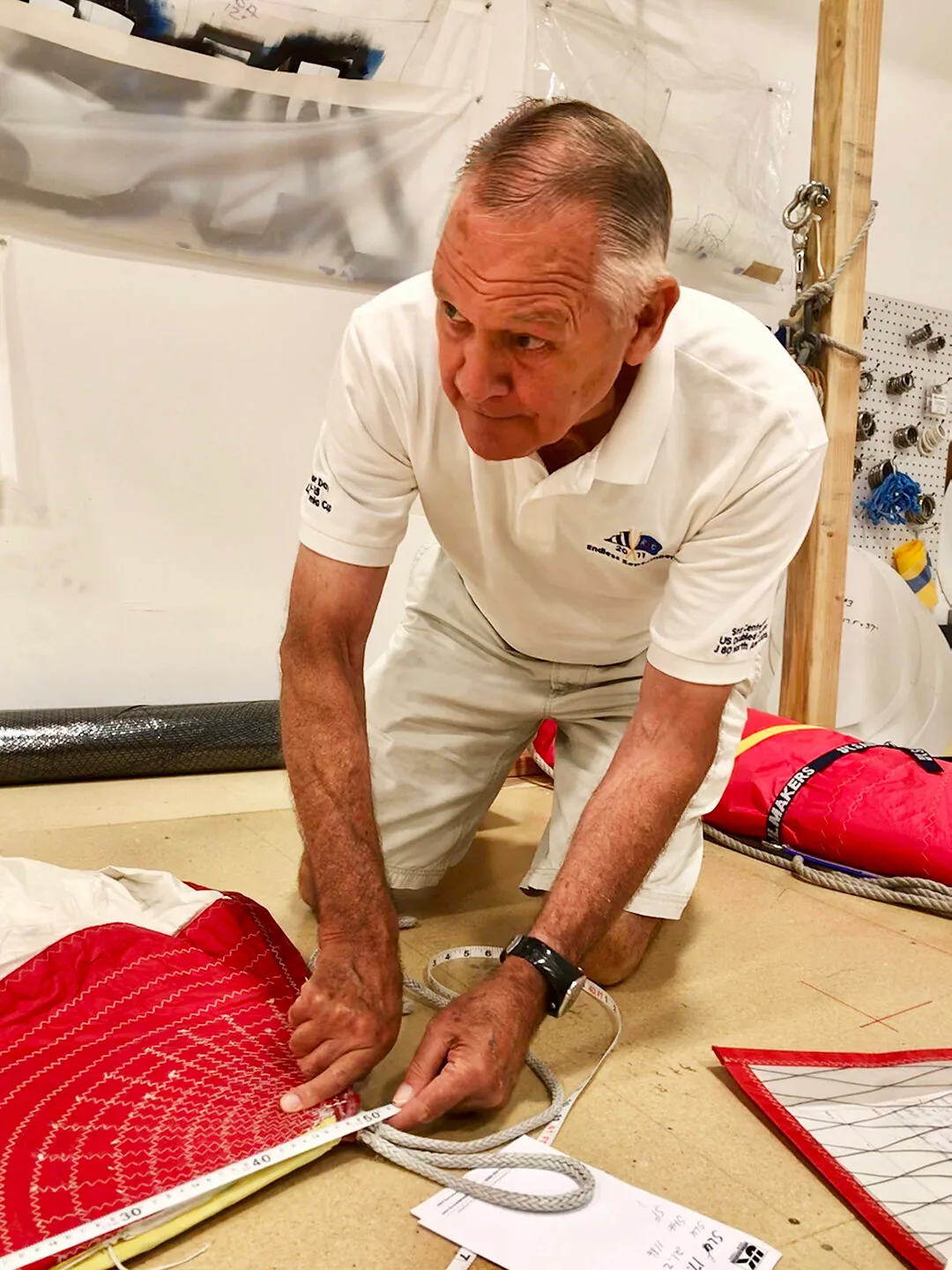 Grey-haired sailmaker in a white polo kneeling on the loft floor, measuring a red sail panel with a tape